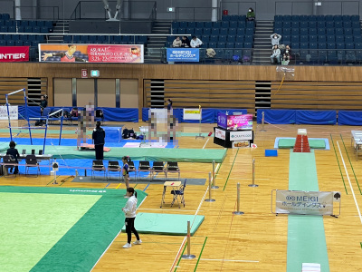 2026年3月21日（土）第3回寺本明日香カップ会場。跳び箱、段違い平行棒、タンブリングバーンが写っています。株式会社MEIGIホールディングスの社旗が２枚写っています。